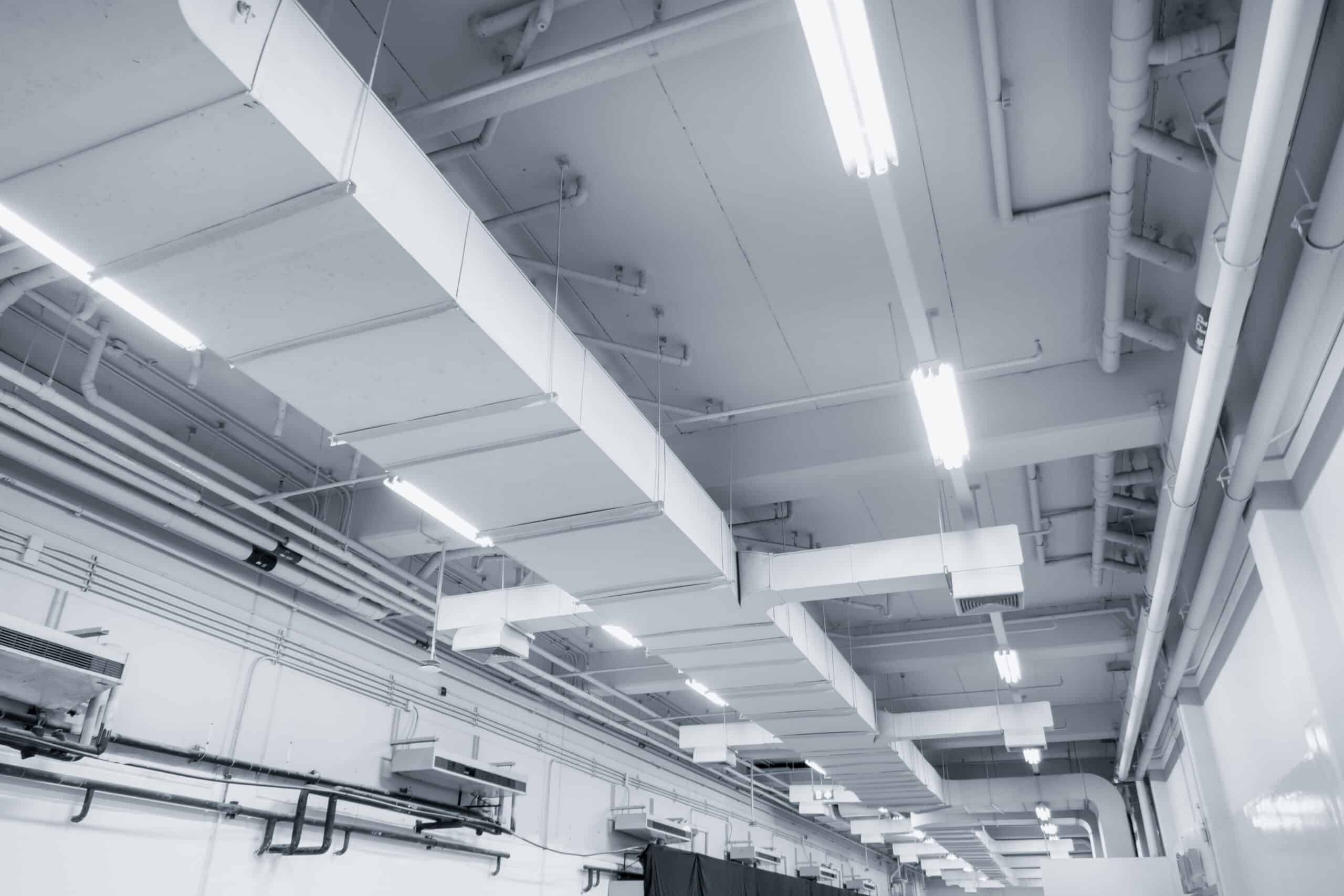 Intérieur industriel ou entrepôt avec conduits de ventilation rectangulaires apparents, tuyauterie et éclairage fluorescent blanc froid.