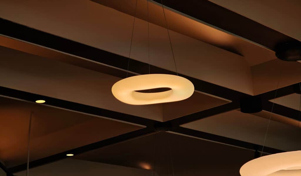 Lustre Design Torsadé Moderne Plafond Éclairage Lustre design torsadé jaune pâle suspendu sous un plafond moderne marron avec des poutres noires et des spots encastrés.