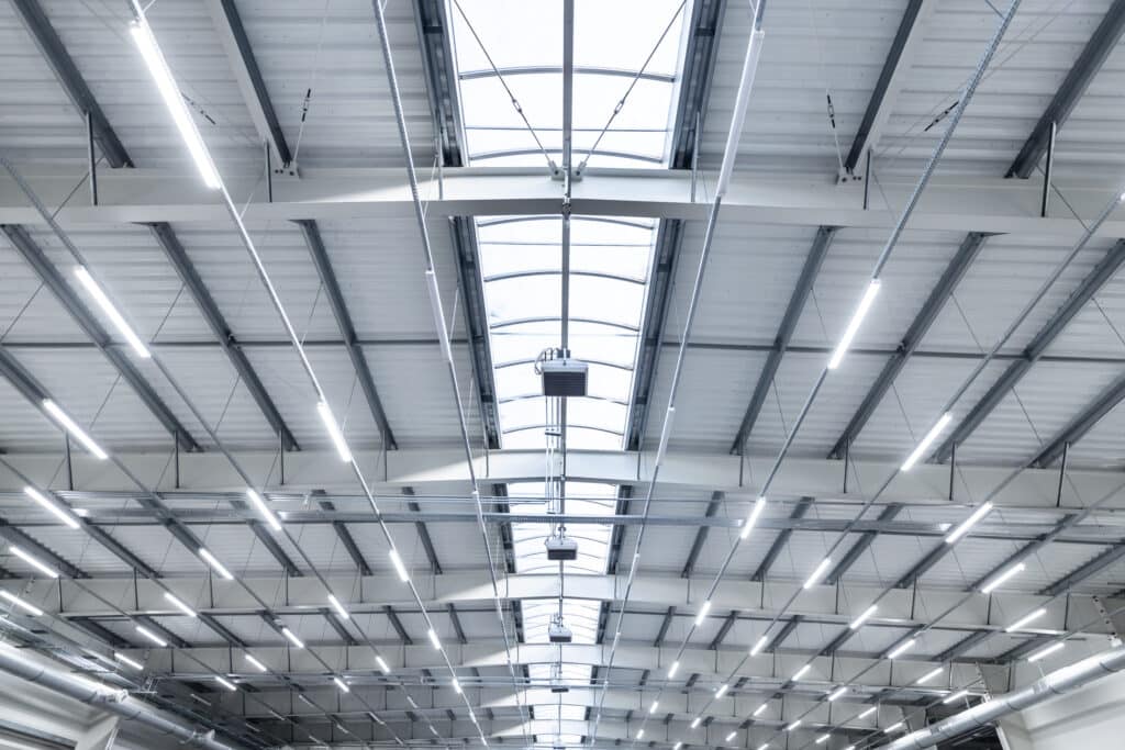 Plafond entrepôt moderne : structure, éclairage et puits de lumière Intérieur d'un grand entrepôt industriel moderne avec toit métallique, puits de lumière zénithale et longs luminaires LED blancs.