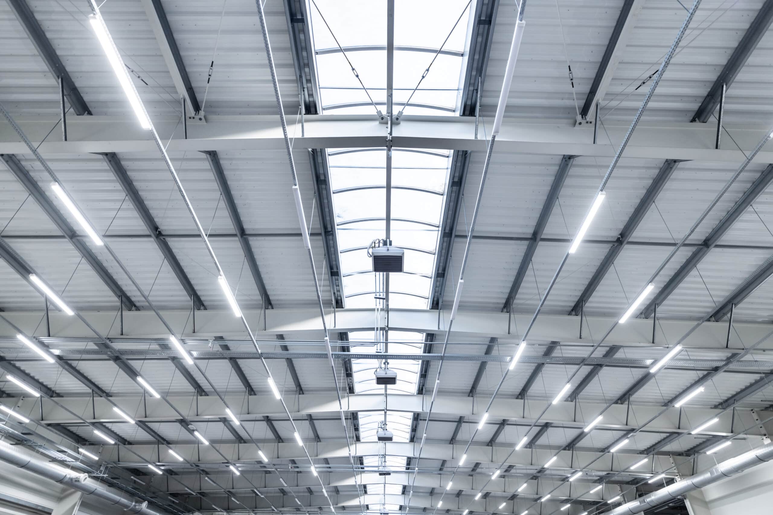 Intérieur d'un grand entrepôt industriel moderne avec toit métallique, puits de lumière zénithale et longs luminaires LED blancs.
