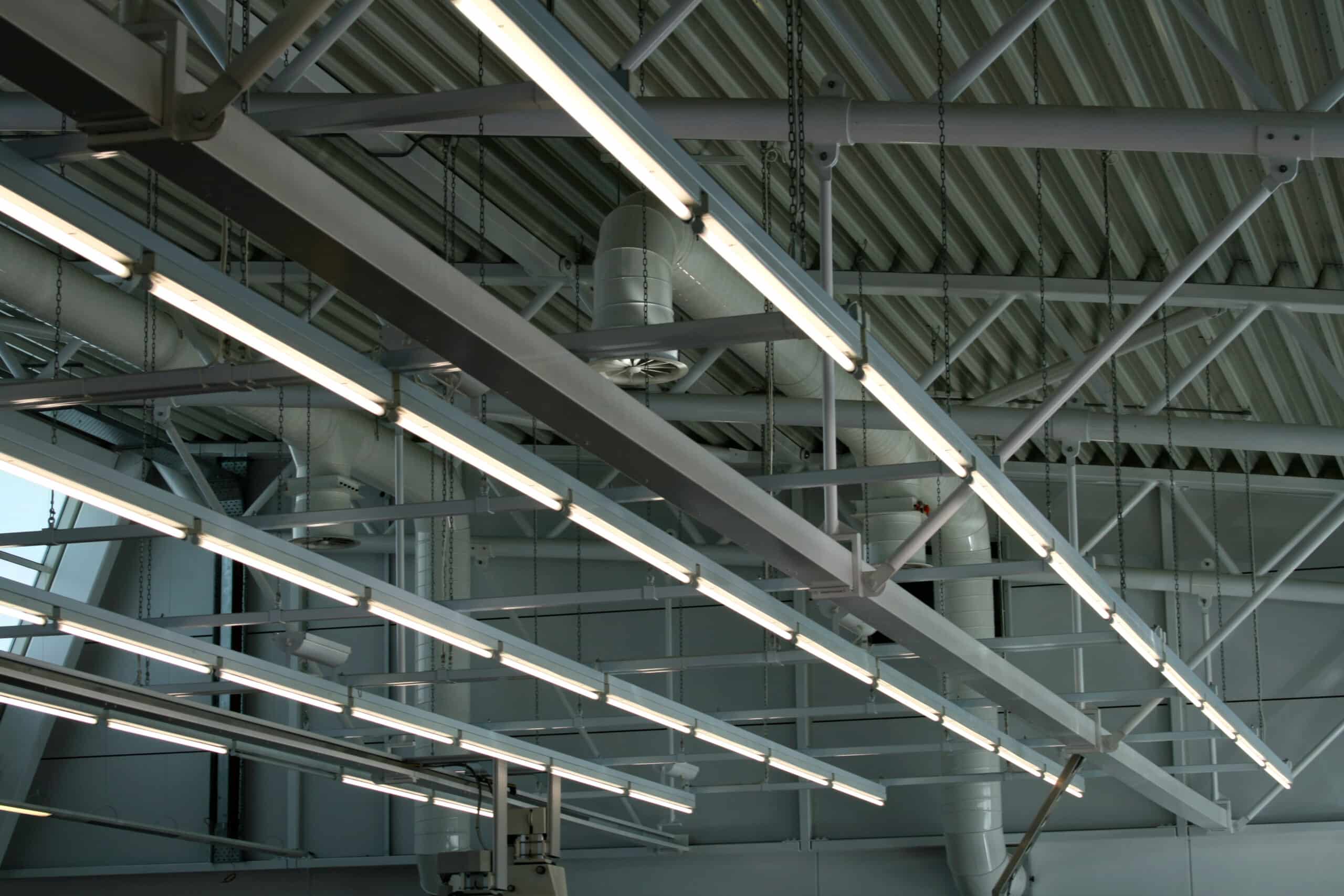 Plafond industriel avec poutres métalliques, néons lumineux suspendus par chaînes et conduits de ventilation blancs.