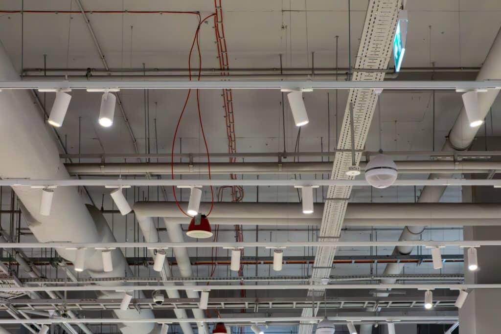 Plafond technique commerce : éclairage et gaines apparentes Plafond technique d'un commerce avec conduits d'aération blancs, chemins de câbles perforés, projecteurs orientables blancs et un panneau de sortie de secours vert.