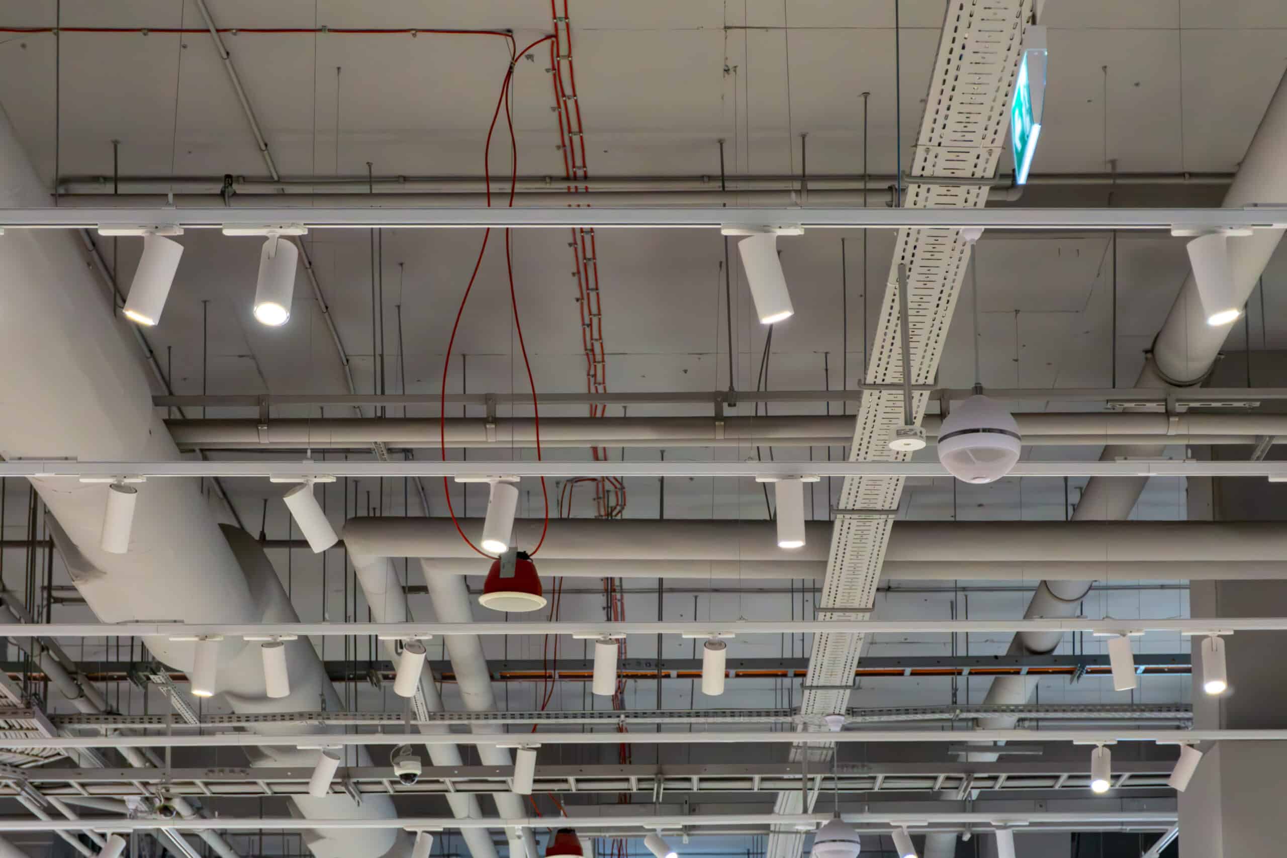 Plafond technique d'un commerce avec conduits d'aération blancs, chemins de câbles perforés, projecteurs orientables blancs et un panneau de sortie de secours vert.